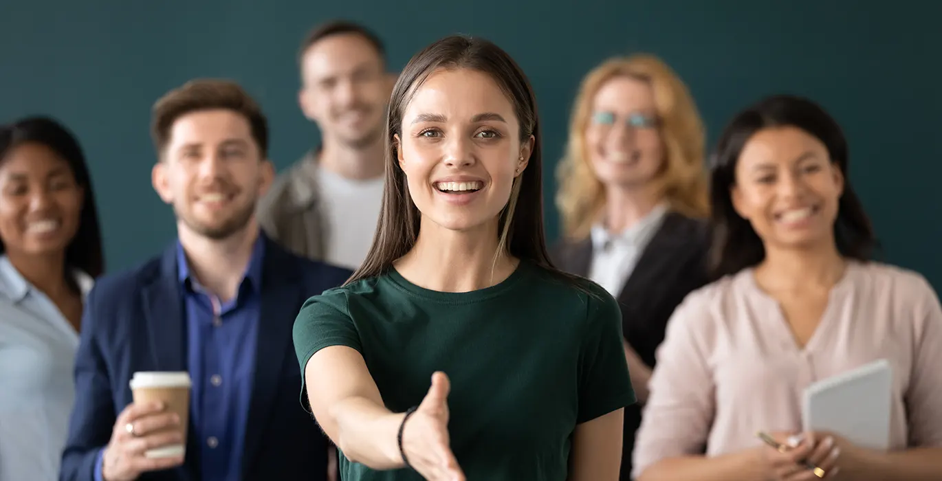 Group of people in business attire. The person at the front is holding their hand out toward the camera for a handshake.
